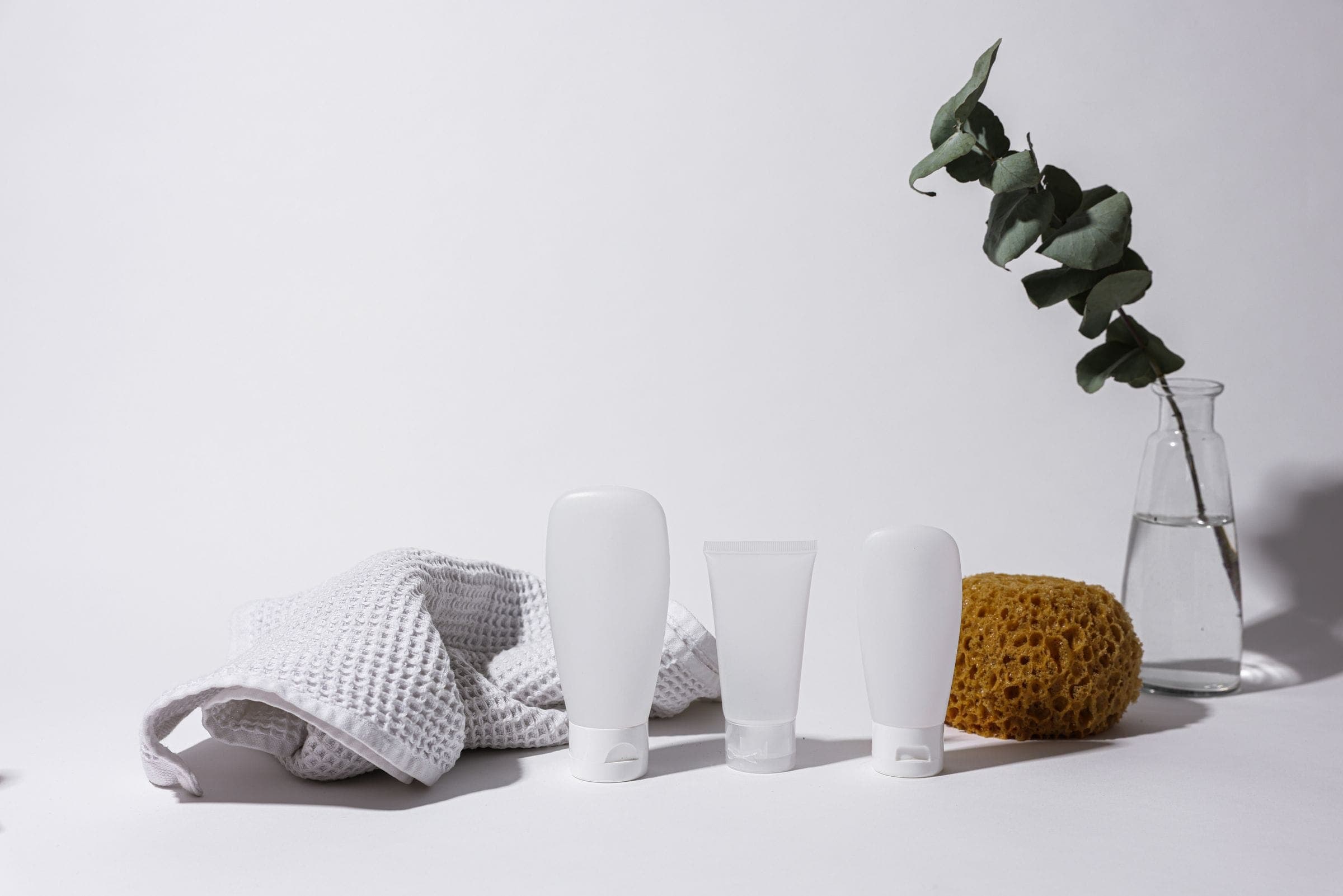 Unbranded white cosmetic bottles, a waffle towel and a sea sponge beside a eucalyptus sprig
